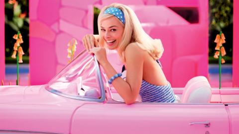 Actor Margot Robbie as Barbie, looking at the camera and smiling as she leans over the steering wheel of her trademark pink convertible.